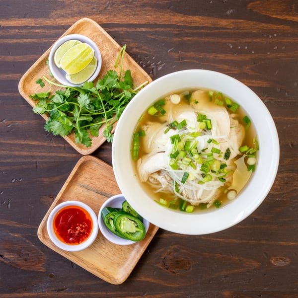 Overhead view of a bowl of Vietnamese chicken pho with lime, cilantro, chili sauce, and jalapeños.
